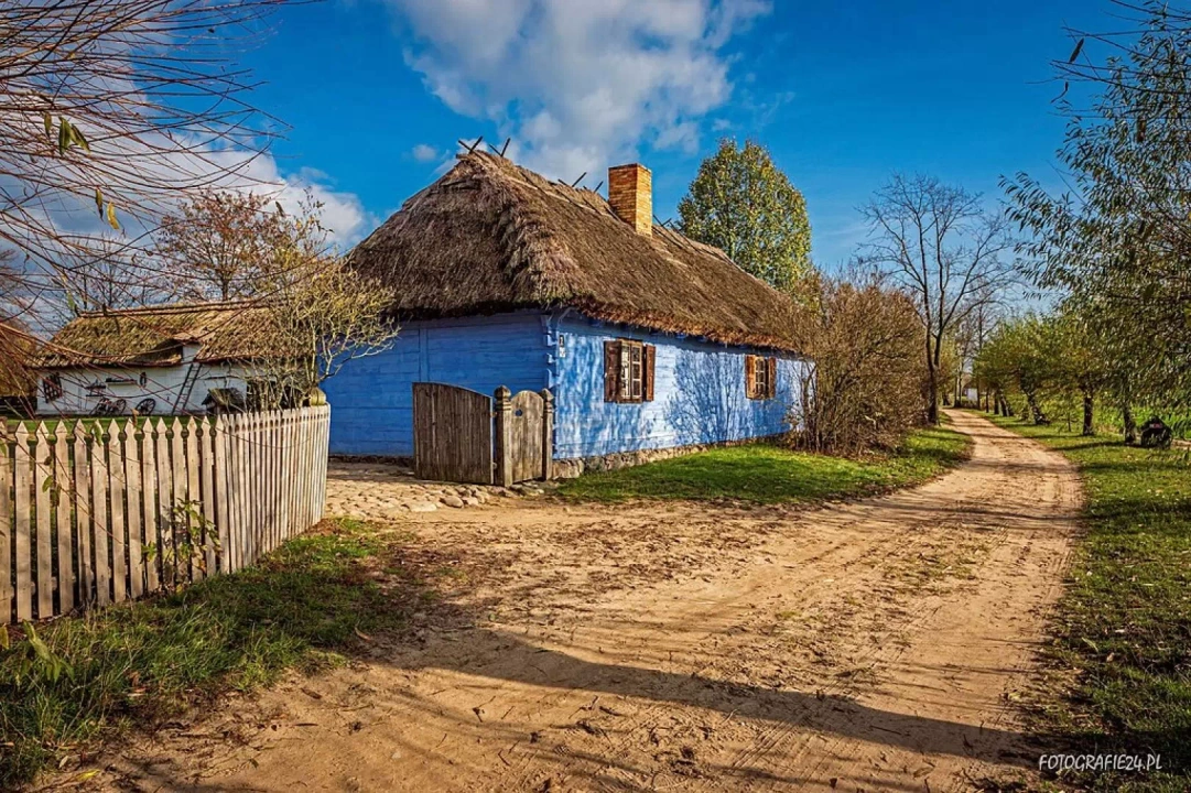 Tradycyjna, drewniana chata kryta strzechą, otoczona drewnianym płotem, z ogrodem pełnym zieleni.