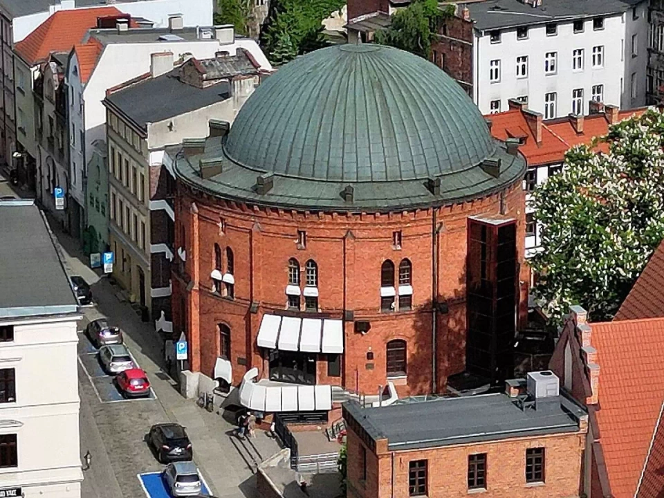Budynek Planetarium w Toruniu. Obiekt z czerwonej cegły w kształcie rotundy otoczony wieloma budynkami starego miasta.