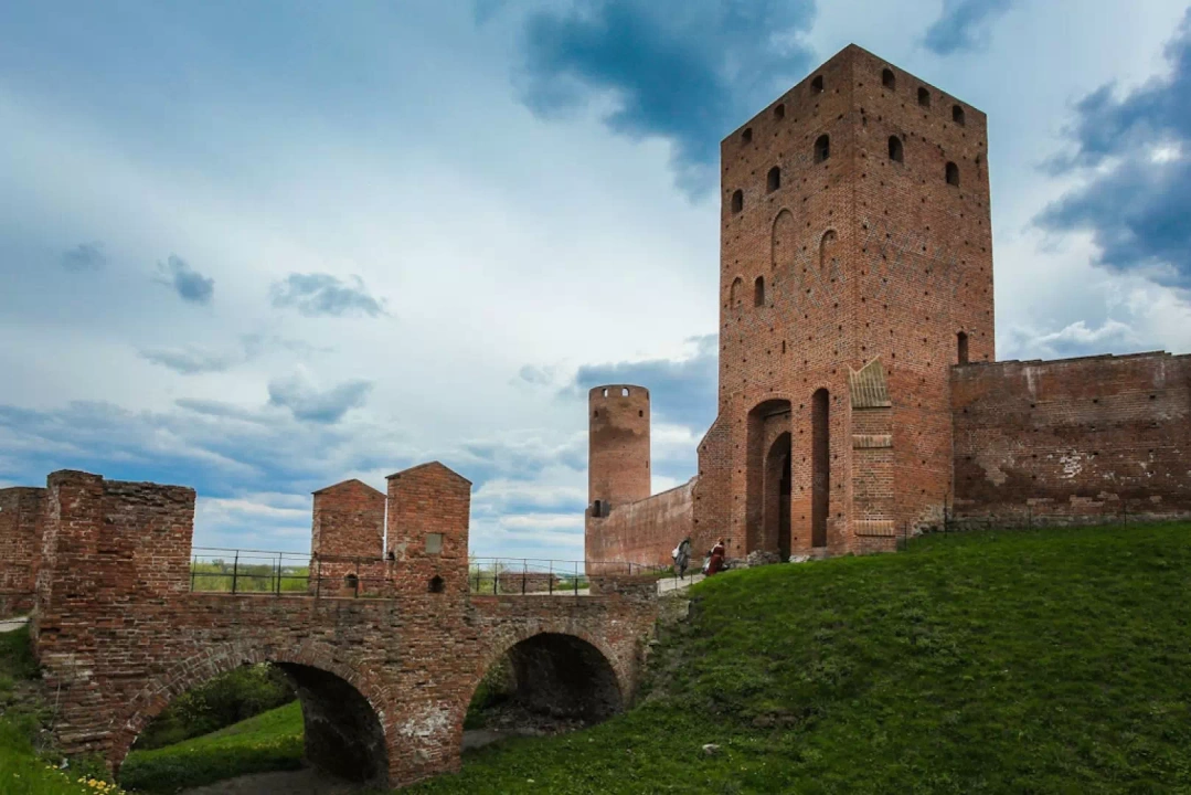 Zamek, a właściwie jego ruiny, o gotyckiej architekturze z wysoką, ceglano-murarską wieżą i mostem łukowym na zielonym terenie. Widok zamku otoczonego murami obronnymi i niebem pełnym chmur.