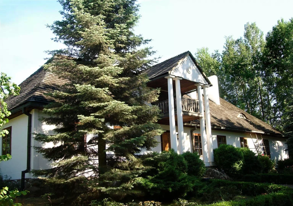 Drewniany wiejski dom kryty strzechą, otoczony zielenią, z kwitnącymi roślinami w przydomowym ogródku. Tradycyjna architektura wiejska, przedstawiająca zabudowę charakterystyczną dla dawnej polskiej wsi.